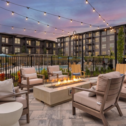Cushioned lounge chairs around a lit fire pit under string lights at Accent Springs, with the pool area behind